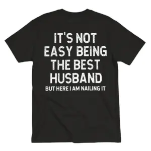 Back flat lay of a black t-shirt reading It's Not Easy Being The Best Husband But Here I Am Nailing It in white text.