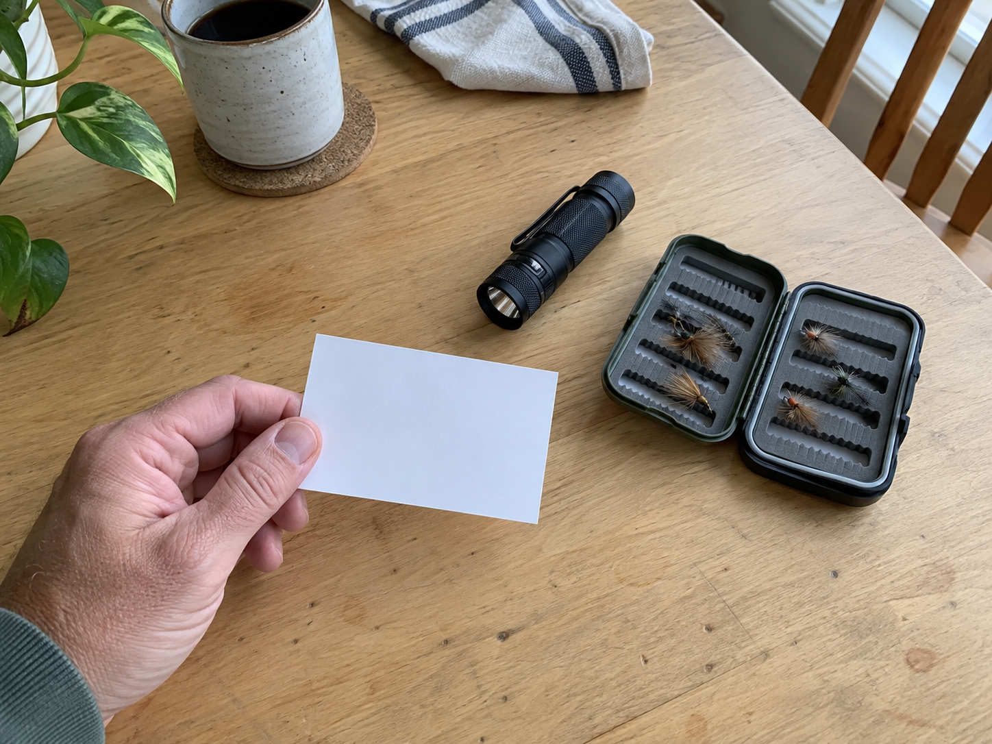 Gifts for Plumbers: Practical Picks They’ll Appreciate 2 A person holds a blank gift card beside a flashlight and an open case of fishing flies on a table.