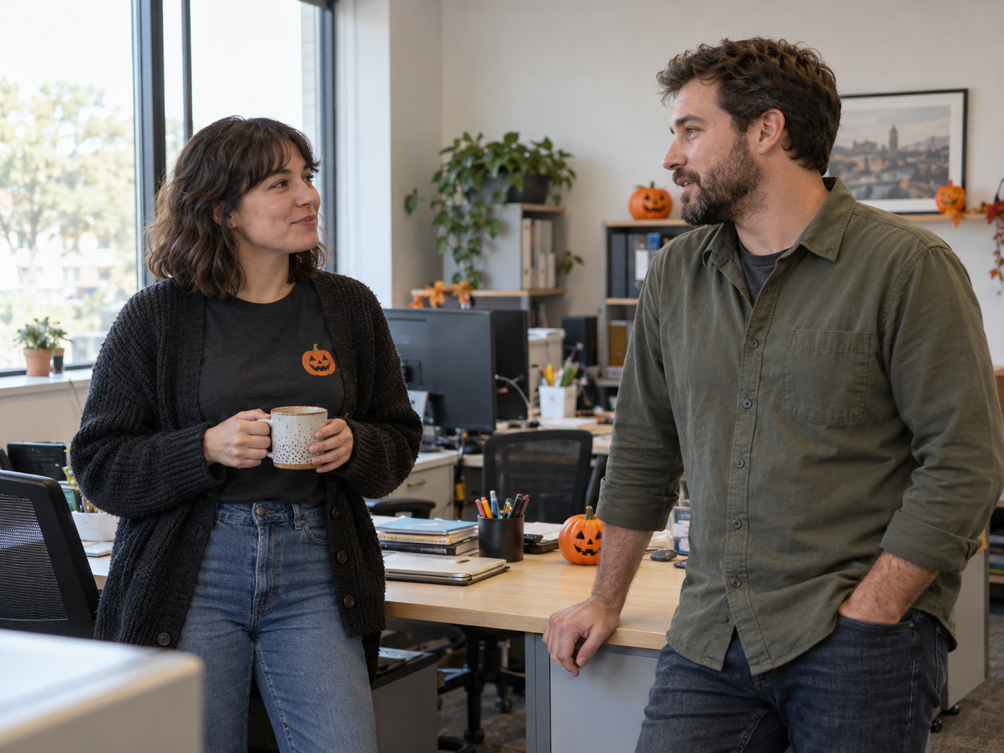 Funny Halloween Shirt Ideas: Picking the Right Joke for Your Event 2 Two coworkers chatting in an office, with one wearing a charcoal pumpkin graphic tee under an open dark cardigan.