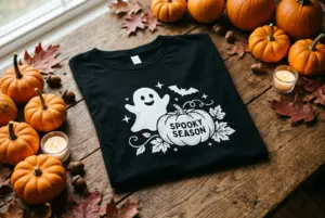 Folded black graphic Halloween T-shirt with a white ghost and pumpkin design reading Spooky Season laid on a rustic wooden table with mini pumpkins, fallen autumn leaves, and candles.