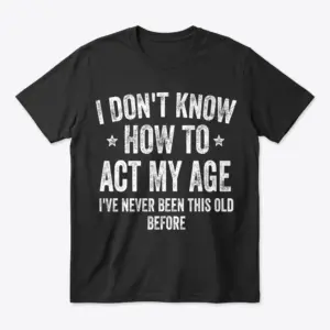 Flatlay of a black t-shirt printed with distressed text reading 'I Don't Know How to Act My Age I've Never Been This Old Before'.