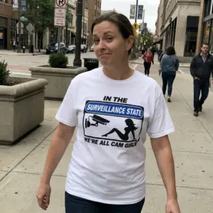 In The Surveillance State We're All Cam Girls Funny T-Shirt 10 Everyday streetwear outfit styling white shirt with white tee front print reading IN THE SURVEILLANCE STATE WERE ALL CAM GIRLS with a CCTV sign