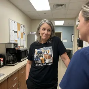Literally Just An LPN Raccoon Iced Coffee Funny Nurse T-Shirt 11 Relaxed hospital breakroom scene wearing black tee with black tee front print of a raccoon in blue scrubs, LPN cap, sunglasses, and stethoscope drinking