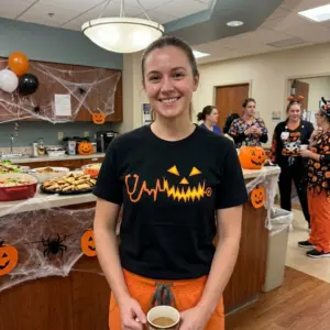 Nurse Stethoscope Heartbeat Glowing Pumpkin Halloween T-Shirt 11 Orange Halloween stethoscope and heartbeat pumpkin graphic on a black T-shirt, shown on a man in a clinical setting.